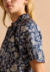 Námornícky modrá kvetinová blúzka so zberaným výstrihom, krátkymi rukávmi a bielym vzorom zobrazením kvetov a listov. Mäkka textúra látky.