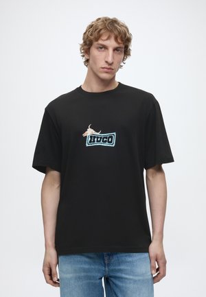 Ung mand med lysbrunt hår iført en sort T-shirt med et blåt "HUGO" logo og en tegning af en okseskaldd, parret med lyseblå jeans.