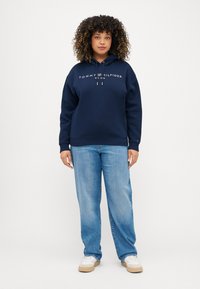 Felpa oversized blu navy con logo frontale, cappuccio con coulisse e polsini a costine, abbinata a jeans a gamba dritta azzurri e sneakers bianche.