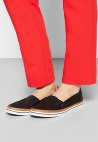 Pieds portant des baskets noires sans lacets avec une bordure marron et des semelles blanches, associées à un pantalon rouge vif sur fond gris.
