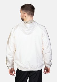 Chaqueta blanca ligera con capucha, con detalles en beige en los hombros y los puños. Cintura elástica y tejido texturizado.