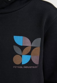 Zwarte sweatshirt met een logo van blauwe, bruine en grijze geometrische vormen. Onder het ontwerp staat de tekst "PETROL INDUSTRIES". Zachte textuur.