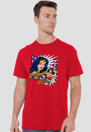 Mann trägt ein rotes T-Shirt mit einer bunten Vintage-Wonder-Woman-Grafik und Text auf der Vorderseite vor einem schlichten grauen Hintergrund.