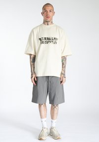 T-shirt oversize crema con grafica nera "MULTIPLY APPAREL", abbinato a pantaloni grigi, calze bianche e sneakers beige. Sfondo semplice.
