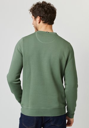 Homme portant un t-shirt vert uni à manches longues, vu de dos, avec des cheveux bouclés foncés et un jean bleu foncé sur un fond clair uni.