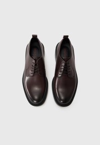 Chaussures en cuir marron foncé avec une surface lisse, bout rond, laçage à l'avant, et semelle en caoutchouc noir avec un motif texturé. Logo visible à l'intérieur.