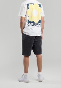 Henkilö, joka on pukeutunut valkoiseen T-paitaan, jossa on suuri keltainen kukka ja "CALIFORNIA"-teksti, mustiin denim-shortsit, valkoiset sukat ja valkoiset lenkkarit.