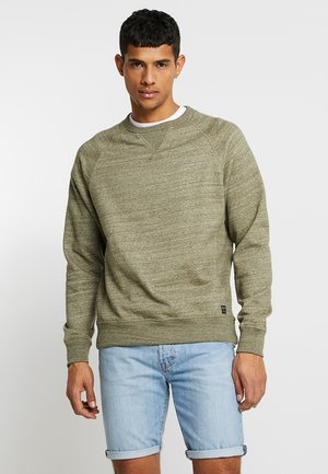 Sweatshirt vert olive en tissu texturé, col rond et poignets côtelés. Porté avec un short en denim bleu clair, arborant une petite étiquette de marque.