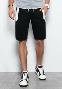 Pantaloni sportivi neri con tasche accentate bianche, vita regolabile con stringa e una vestibilità comoda, abbinati a scarpe da ginnastica nero e bianco.