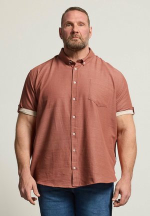 Stämmiger Mann mit kurzen Haaren und Bart, der ein rostfarbenes kurzärmeliges Hemd mit Knöpfen und blaue Jeans trägt, steht vor einem schlichten Hintergrund.
