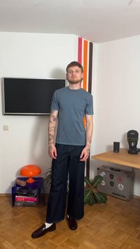 Chemise gris à manches courtes associée à un jean large sombre et des mocassins marron. La pièce comprend une lampe orange et un mur à rayures colorées.