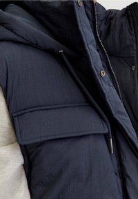 Gros plan d'une veste matelassée bleu foncé avec capuche, grande poche à rabat, fermeture éclair et détail lacet portée sur une manche gris clair.