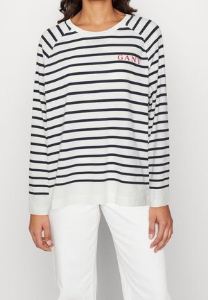 Kvinde iført hvid langærmet top med sorte horisontale striber og rød "GANT"-logo, kombineret med hvide bukser, stående foran en ensfarvet baggrund.