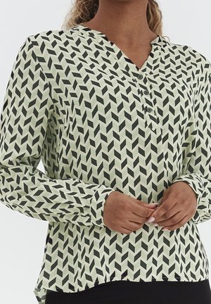 Femme portant un chemisier à manches longues à motifs avec des formes géométriques noires et des poignets boutonnés, mains jointes près de la taille.