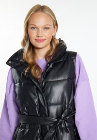 Schwarze Steppweste mit hohem Kragen und Gurtbund, aus glänzendem Kunstledermaterial und mit einem Reißverschluss vorne. Trägt eine lila Bluse.