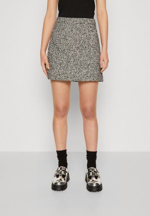 Vrouw met een zwart-witte, getextureerde minirok, zwarte sokken, metalen zilveren stevige schoenen en een zwarte top tegen een effen lichte achtergrond.