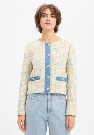 Vrouw draagt een textuurrijke crèmekleurige en blauwe tweedjas met gouden knopen en denim randen, gecombineerd met een lichtblauwe high-waist spijkerbroek.