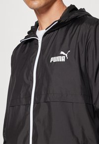 Chaqueta cortavientos negra hecha de material ligero, con un zipper blanco, capucha de malla y logo de Puma en el pecho.