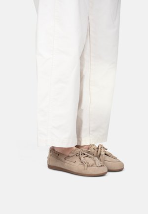 Beige Wildleder-Mokassins mit Quasten, getragen zu locker sitzenden weißen Hosen, gezeigt vom Unterschenkel abwärts vor weißem Hintergrund.