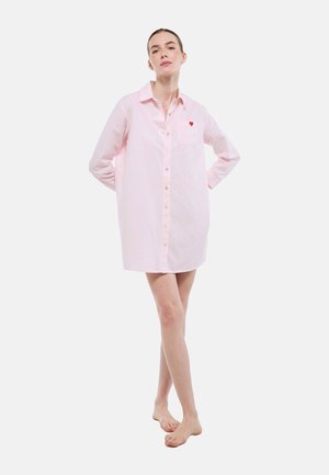 Femme debout pieds nus portant une chemise oversize rayée rose clair avec un petit cœur rouge sur la poche poitrine.