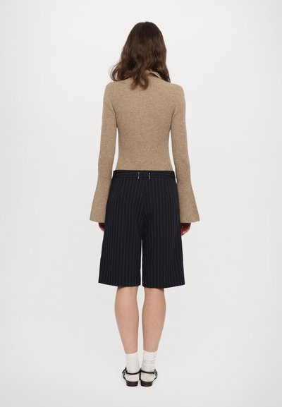 Beige ribbet genser med flagrende ermer kombinert med marineblå rutete shorts. Outfiten har en tettsittende midje og en løs ben silhuett.