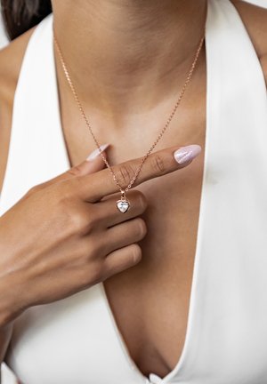 Rose gold necklace featuring a heart-shaped pendant with a clear gemstone, worn on bare skin with a white garment. Hand gesture highlighting the piece.
