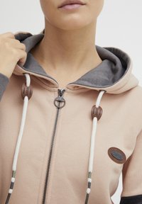 Parka s kapuljačom u svijetlo ružičastoj boji s sivim detaljima, s prednjim zatvaračem, tkanim zatvaračima i logom na lijevoj strani prsiju.
