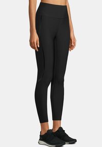 Czarne legginsy o wysokim stanie wykonane z elastycznego materiału, z eleganckim fasonem, bocznymi kieszeniami i kontrastowymi szwami. Połączone z czarnymi trampkami.