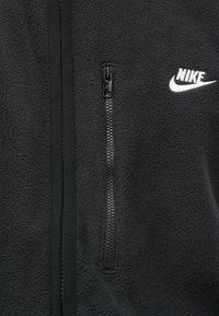 Jachetă din fleece neagră cu o textură netedă, dotată cu un buzunar cu fermoar și un logo alb Nike pe piept. Închidere cu fermoar pe toată fața.
