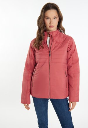 Kvinde iført en koralfarvet, quiltet jakke med frontlynlås og lynlåslommer, kombineret med blå jeans, står mod en hvid baggrund.