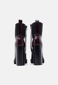Ζευγάρι γυαλιστερές σκούρες μπορντό μποτάκια μέχρι τον αστράγαλο με χοντρά ψηλά τακούνια και μαύρες ελαστικές πλευρικές επένδυσεις, εμφανιζόμενα από πίσω πάνω σε λευκό φόντο.