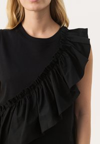 Black top featuring a round neckline and a prominent ruffled panel on one side, made of soft fabric with gathered texture.