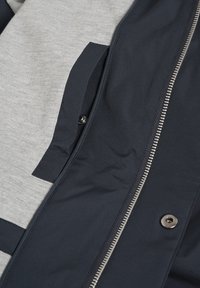 Close-up of a navy jacket with zipper and snap button, layered over a light gray fabric with a navy rectangular patch.
