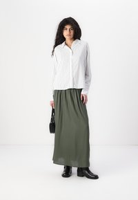 Chemise blanche à rayures, longue jupe verte fluide, bottines noires à la cheville et petit sac à main noir, sur fond blanc uni.