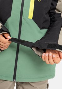 Groene en zwarte waterdichte jas met een gele rits, verstelbare manchetten en een ritssluiting. Kenmerkt zich door een gladde textuur en duurzame stof.