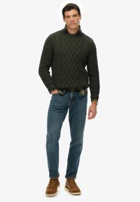 Ciemnozielony sweter z dzianiny z splotem warkoczowym, połączony z koszulą w kratę. Zestawiony z niebieskimi dżinsami i brązowymi butami na sznurówki.
