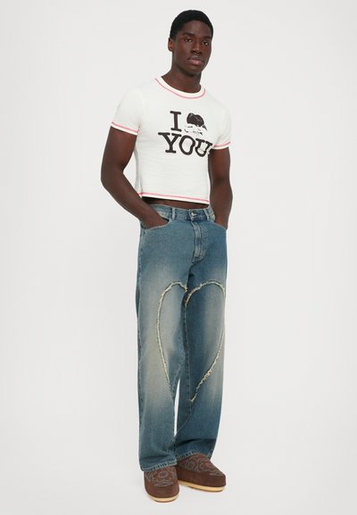 Man staat met zijn handen in zijn zakken, gekleed in een witte crop-top t-shirt met "I [heart] YOU" en blauwe jeans met grote rafelige hartontwerpen op de bovenbenen.