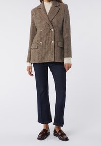 Femme portant un blazer marron texturé à double boutonnage, un pull à col roulé crème, un jean court foncé et des mocassins marron foncé avec un détail chaîne dorée.