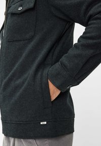 Donkergrijze jas van zacht, gestructureerd stof. Voorzien van een ritsluiting, een knoopzak en een klein merkteken nabij de zoom.
