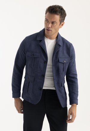 Homme portant une veste bleu marine boutonnée sur un t-shirt blanc, regardant vers sa gauche sur un fond blanc.