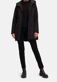 Černá nepromokavá parka s předním zipem, nastavitelnou kapucí a dvěma předními kapsami; nošená přes černý top a kalhoty, doplněná černými botami.