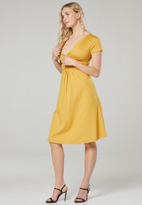 Gelbes, knielanges Kleid mit tailliertem Oberteil, kurzen Ärmeln und gerafftem Taillendetail. Glattes Material und sanfte A-Linie.