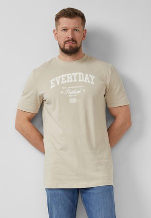 Mężczyzna z brodą, ubrany w beżowy t-shirt z nadrukiem "EVERYDAY Rethink" i niebieskie dżinsy, stoi z rękami za plecami przy szarej ścianie.