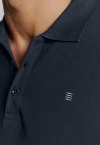 Donkerblauwe polo met een drieknopssluiting, gestructureerde stof en een klein grijs logo nabij de zoom.