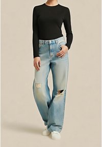 Personne portant un haut noir à manches longues, un jean large déchiré bleu clair et des baskets blanches, debout avec une main dans la poche.