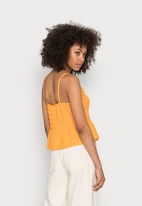 Top peplum laranja com textura franzida, alças finas e corpete ajustado. Usado com calças brancas de perna larga e de cintura alta.