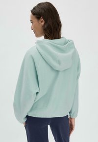Mintgrüner Fleece-Hoodie mit lockerem Schnitt, ausgestattet mit einer geräumigen Kapuze und geripptem Saum. Der Stoff hat eine weiche, strukturierte Optik.