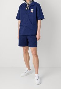 Marineblå kortärmad knapp-up skjorta med patch, matchande shorts och vita sneakers. Slät tyg, avslappnad passform, minimalistisk design.
