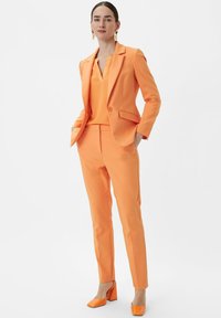 Women's orange suit featuring a fitted blazer, matching top, and slim trousers made of smooth fabric. Paired with orange block-heeled shoes.