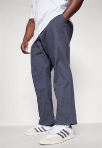 Marineblaue, lockere Chinos aus Baumwollmischung, mit Seitentaschen und einer glatten Textur; getragen mit weißen Sneakers mit grauen Streifen.